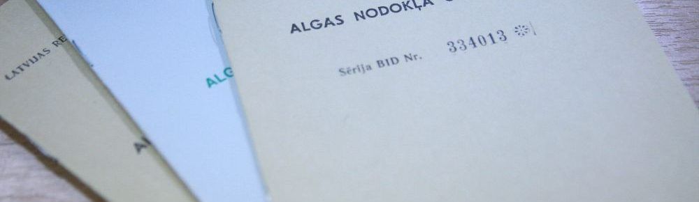 Divu Latvijas algu nodokļu grāmatiņu tuvplāns ar nosaukumu "Algas Nodokļa Grāmatiņa" un uz vāka uzdrukātu kārtas numuru. Grāmatas ir nedaudz izvēdinātas uz koka virsmas. Grāmatas augšdaļa ir smilškrāsas, bet apakšā - zaļa.