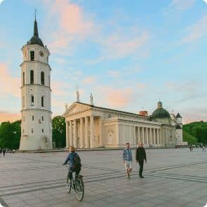 Ainavisks skats uz Viļņas katedrāli un tās zvanu torni Lietuvā. Debesis ir nokrāsotas ar zilām un rozā nokrāsām no rietošās saules. Priekšplānā redzami trīs cilvēki, no kuriem viens brauc ar velosipēdu, bet pārējie divi pastaigājas plašajā laukumā.