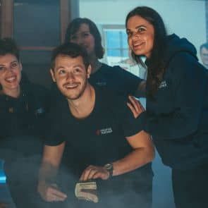 Četri cilvēki, trīs sievietes un viens vīrietis, smaida un pulcējas kopā. Šķiet, ka viņi pozē fotogrāfijai telpās. Vīrietim ir kaut kas nenosakāms. Telpu piepilda viegla migla vai dūmi, un visi valkā ikdienišķu tumšu apģērbu.