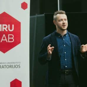 Cilvēks zilā kreklā un tumšā žaketē runā pie izkārtnes ar uzrakstu "MRU LAB". Šķiet, ka fons ir iekštelpu vide, iespējams, universitātē vai laboratorijā. Persona runājot žesti ar rokām.