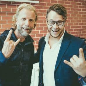 Divi smaidoši vīrieši pozē pie ķieģeļu sienas, katrs rāda žestu “akmens uz rokas”. Vienam ir bārda un tumša jaka ar rāvējslēdzēju, bet otra valkā brilles un tumšu žaketi virs balta krekla.